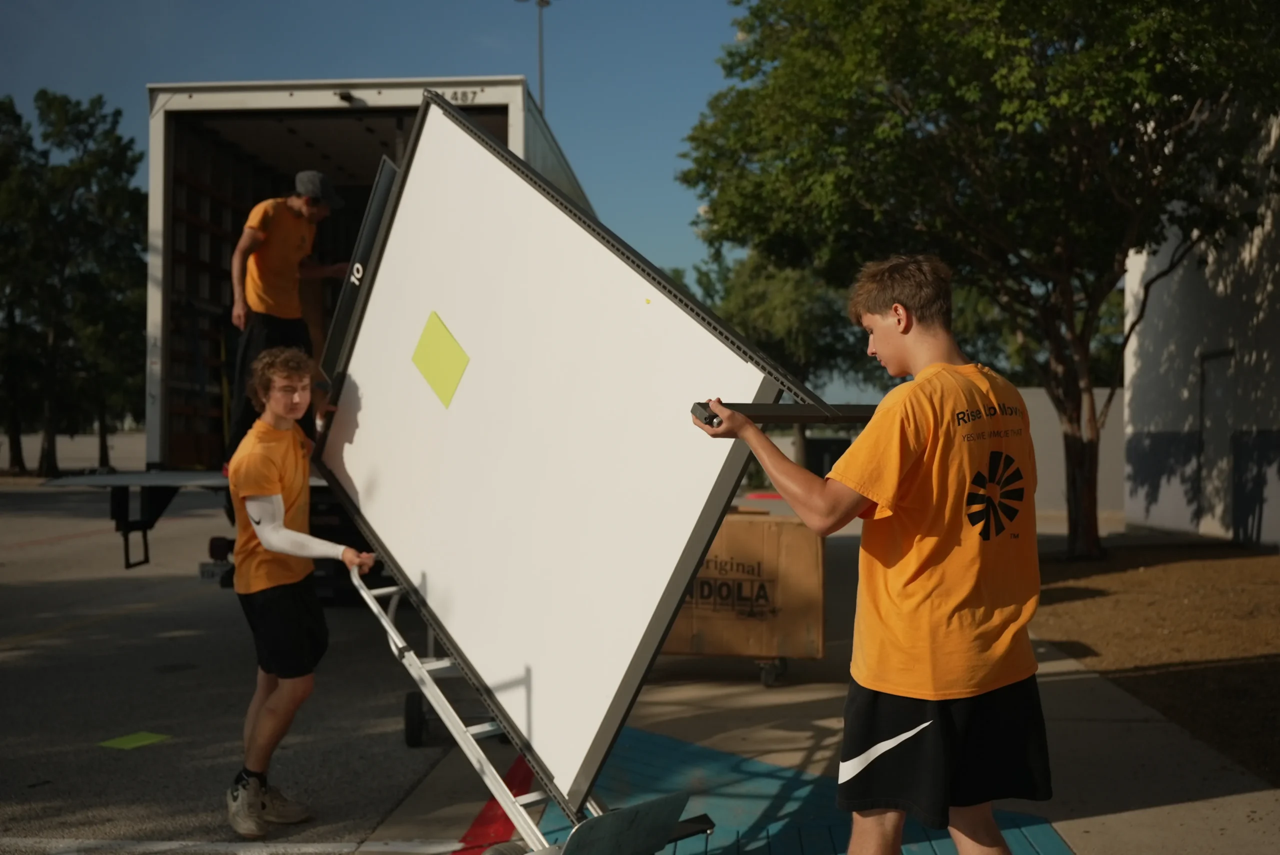 Rise Up Moving movers carrying a large flat panel on a cart toward a moving truck