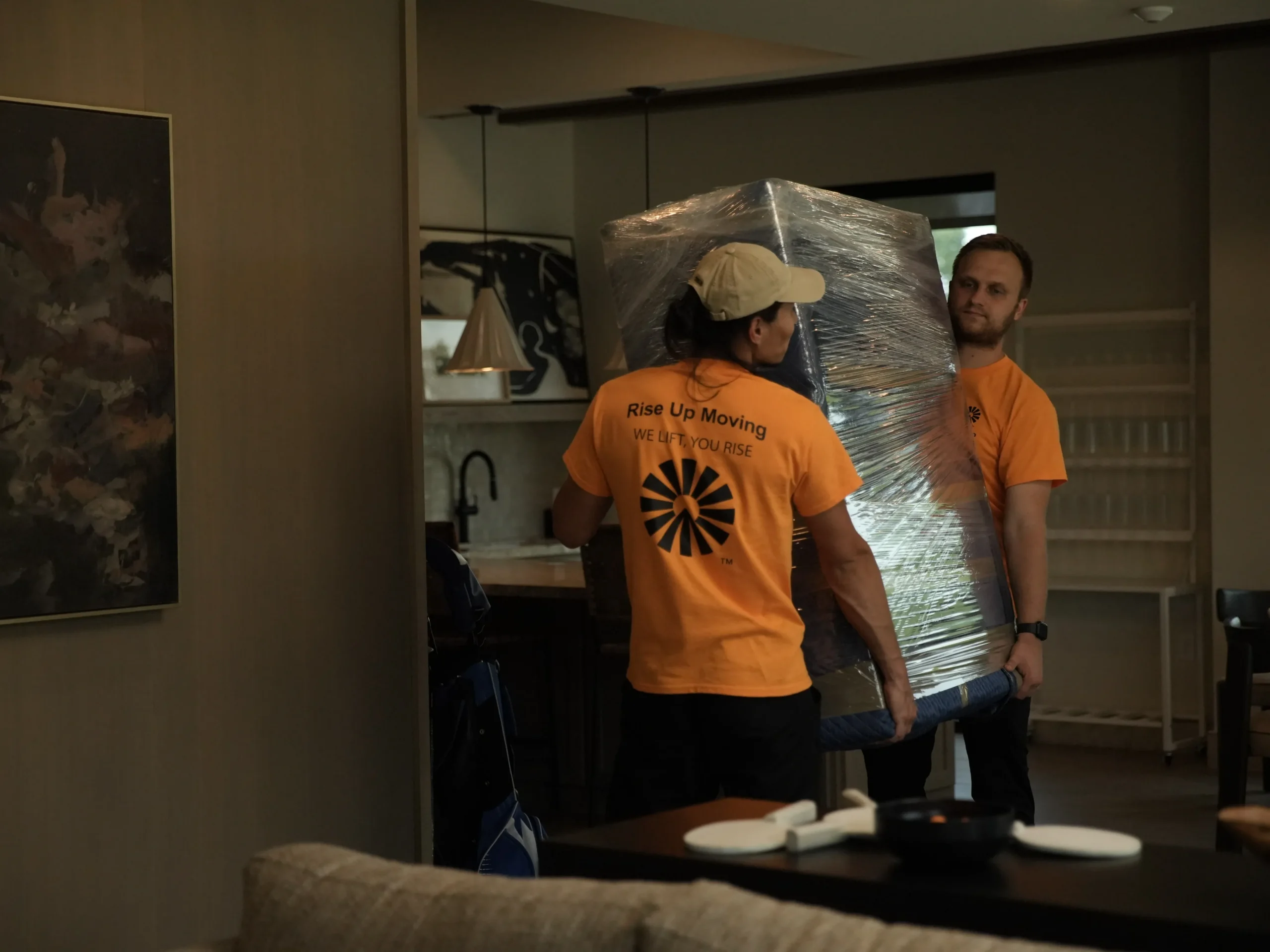 Two Rise Up Moving movers carrying a shrink-wrapped furniture piece through a home